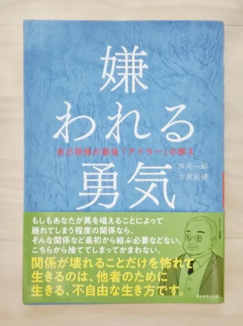 筆者が持っている岸見一郎・古賀史健『嫌われる勇気』の書影。淡い木目の上に置かれた青い表紙と、緑の帯