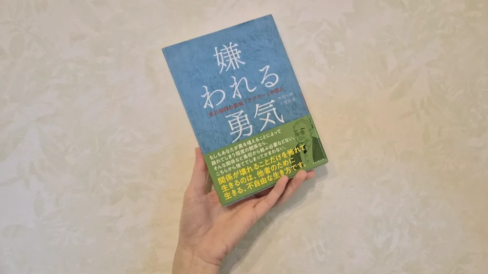 筆者が手に持っている岸見一郎・古賀史健『嫌われる勇気』。青い表紙に緑の帯がかかっている